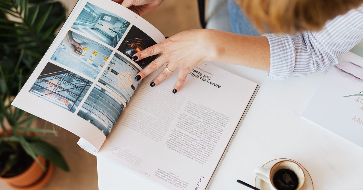 Mãos folheando uma revista sobre a mesa, ao lado de uma xícara de café