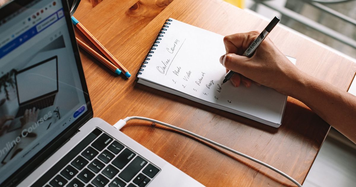 Mão escrevendo anotações num caderno ao lado de um laptop aberto